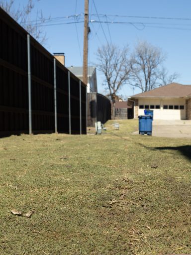 View of a backyard with a wooden fence, grass, and a blue trash bin.