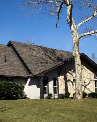 A light-colored house with a sloped roof and a large tree in front on a sunny day.